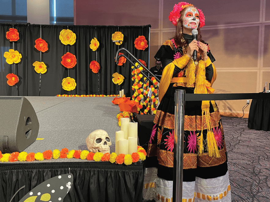 Woman in a traditional Dia de los Muertos dress speaking at an event.