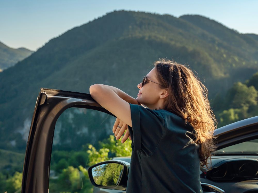 Woman leaning on the open driver side door of a parked car along a mountain road. She is smiling and looking toward a distant mountain peak.